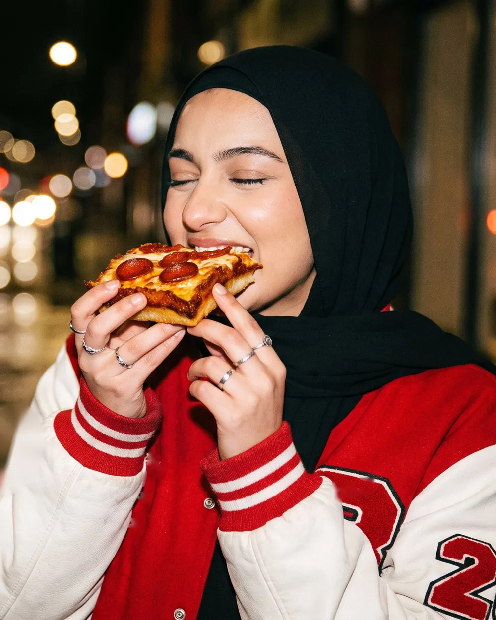 Young woman in a black headscarf eating a Dough Dohs pizza slice on a street at night